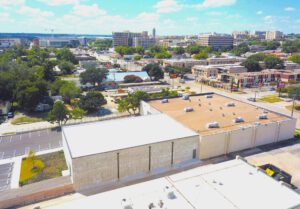 Aerial view of a light grey building with a white and brown roof and views of the city and adjacent street in the background