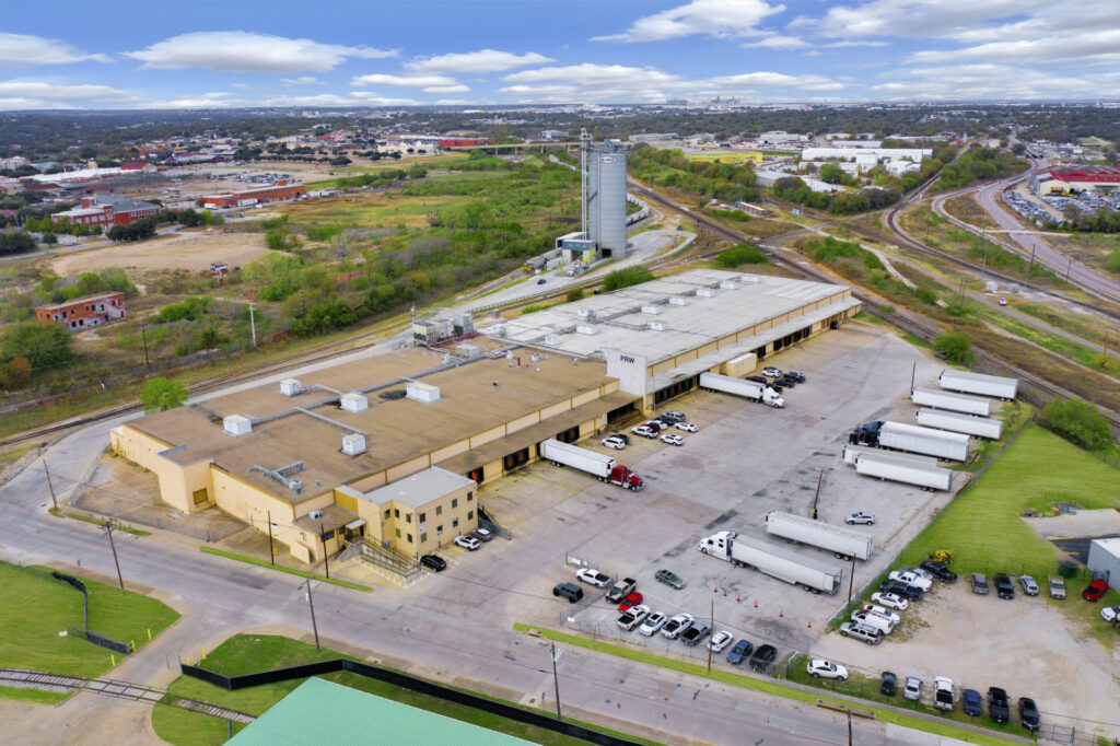 Aerial view of a yellow building with a tan and white roof, multiple loading docks, and a parking lot with cars and trailers