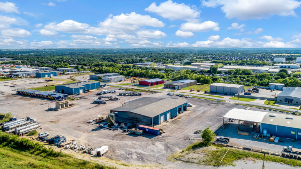 Aerial view of blue industrial buildings with grey roofs with dirt pavement and multiple shipping containers and trailers