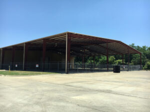 Ground-level view of a large open-air, tin structure with red posts and a barbed wire fence surrounding it