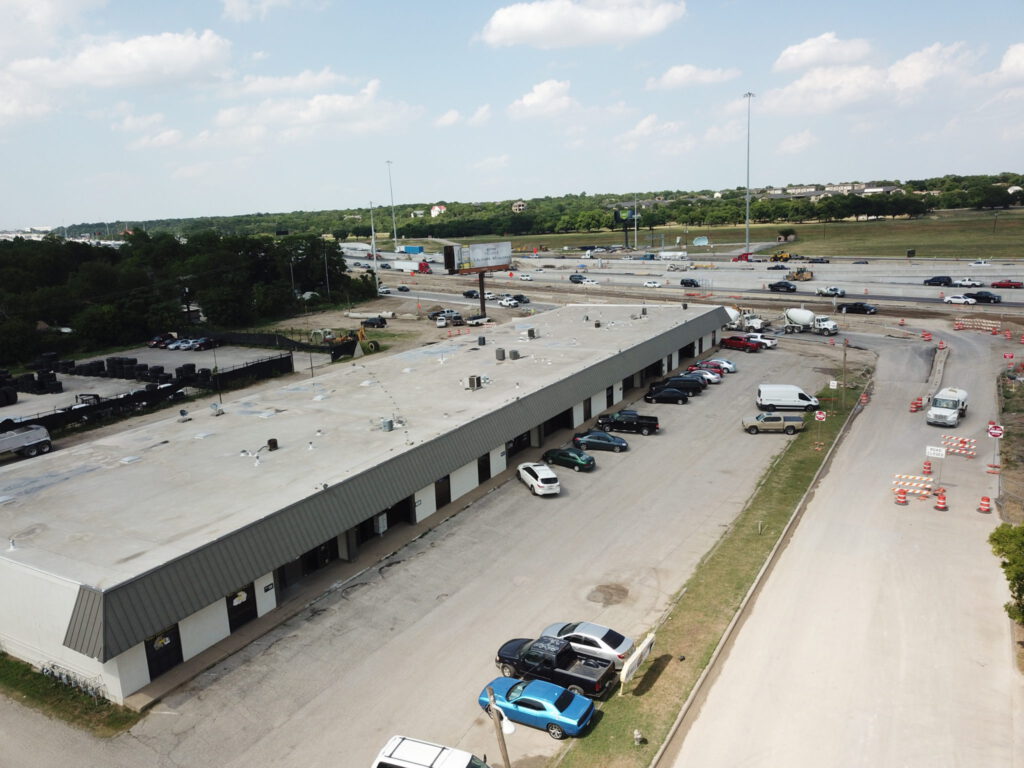 Aerial view of a white building with a grey roof, grey onning, and attached parking lot adjacent to a freeway