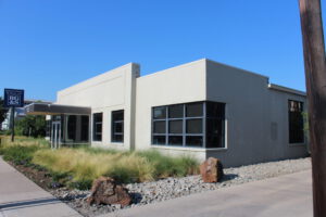 Corner, ground-level view of a grey building with black windows on each side and landscaping with grey and brown rocks